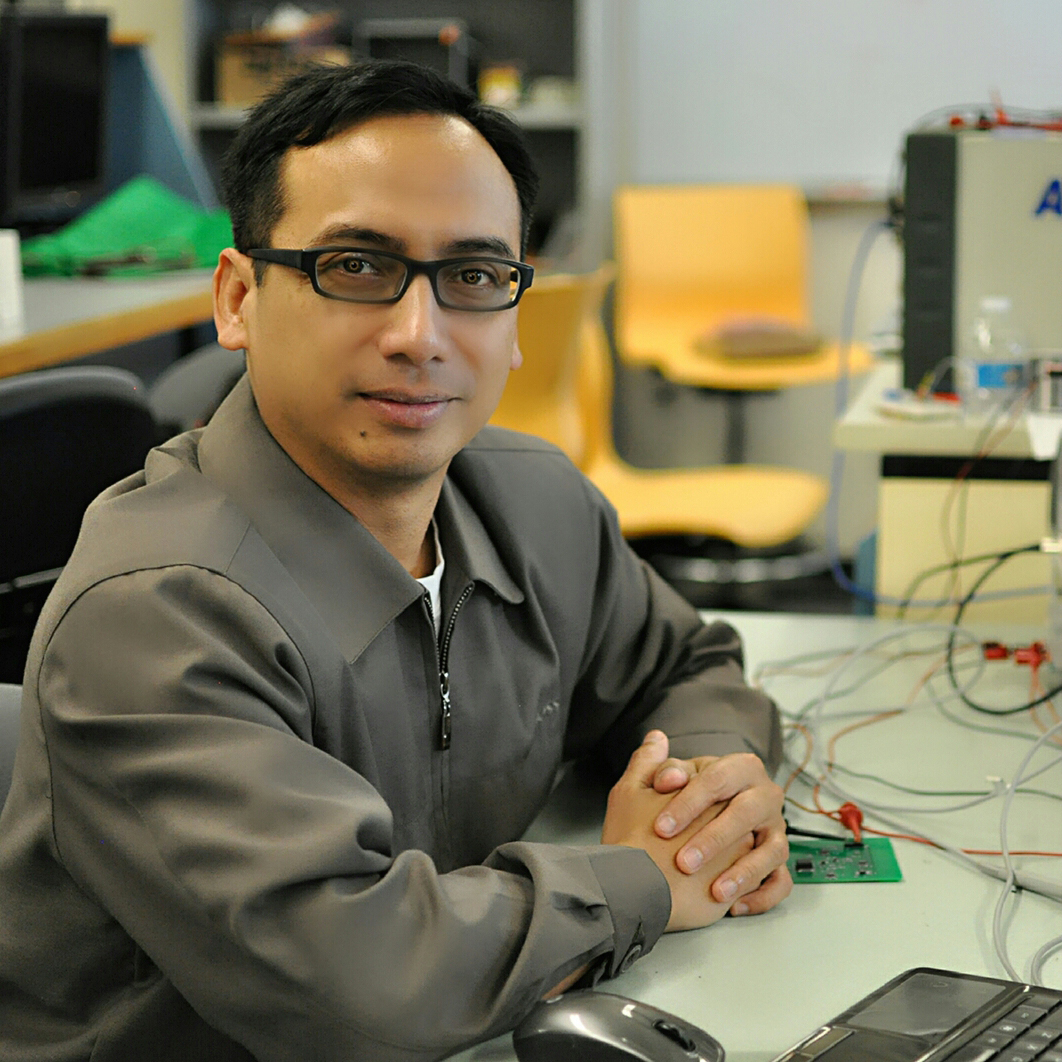 A man in a grey shirt and glasses leans on a desk bearing electrical components