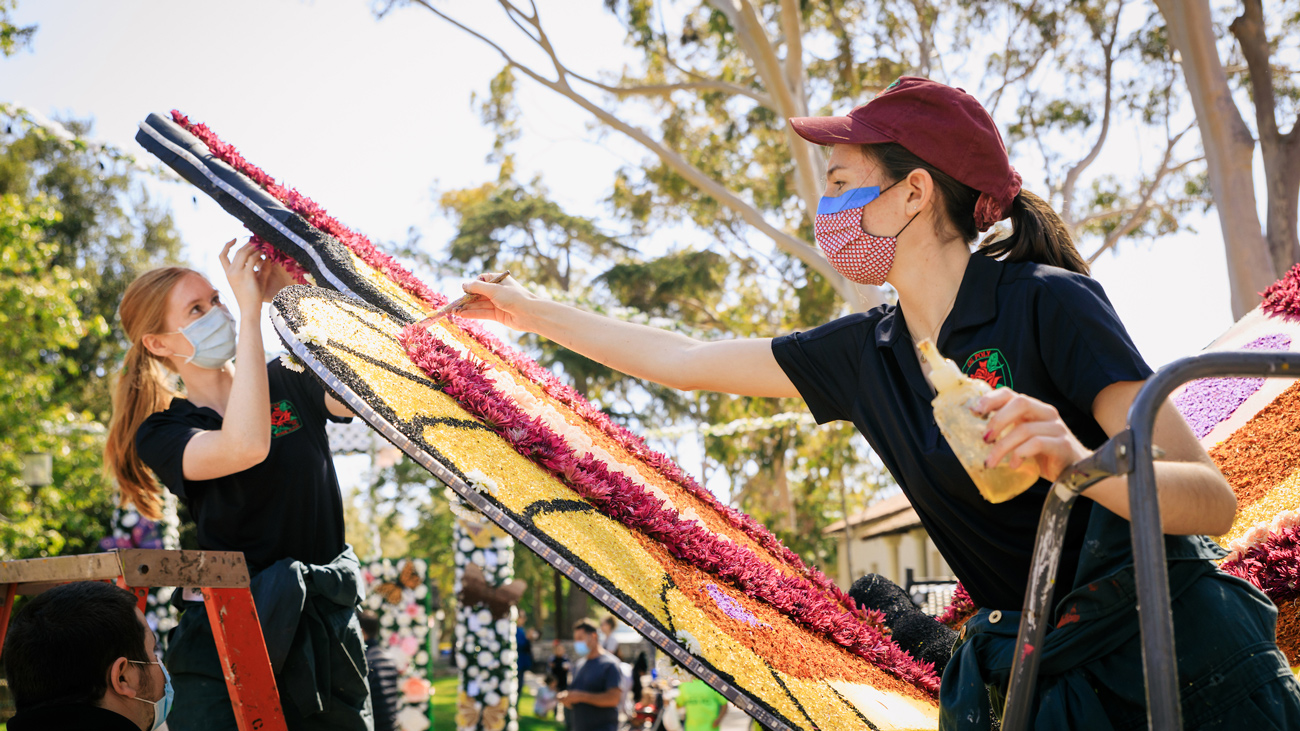 Two students wearing face coverings apply glue and flowers to a butterfly wing installation