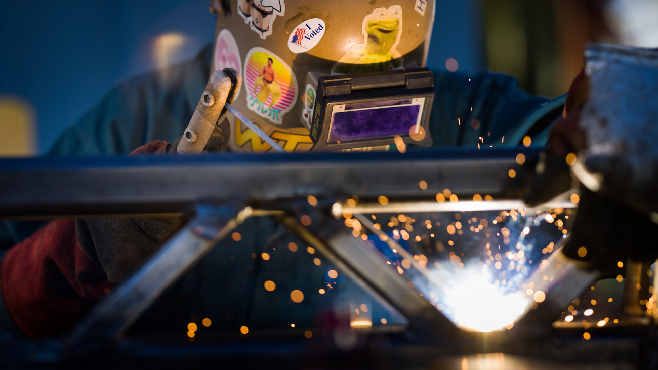 A person in a welding helmet welds metal together with sparks flying in the foreground