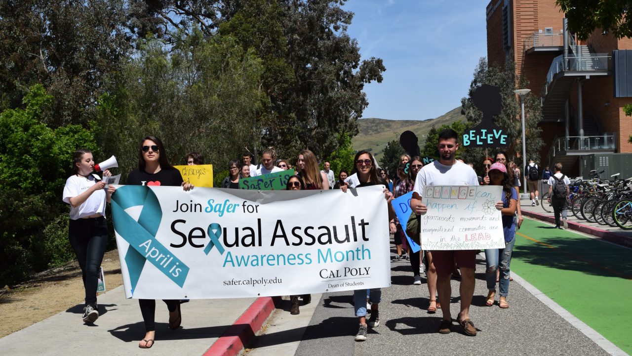 A large group of students march across campus with a banner reading "Join Safer for sexual assault awareness month" and holding other signs about supporting and believing assault survivors