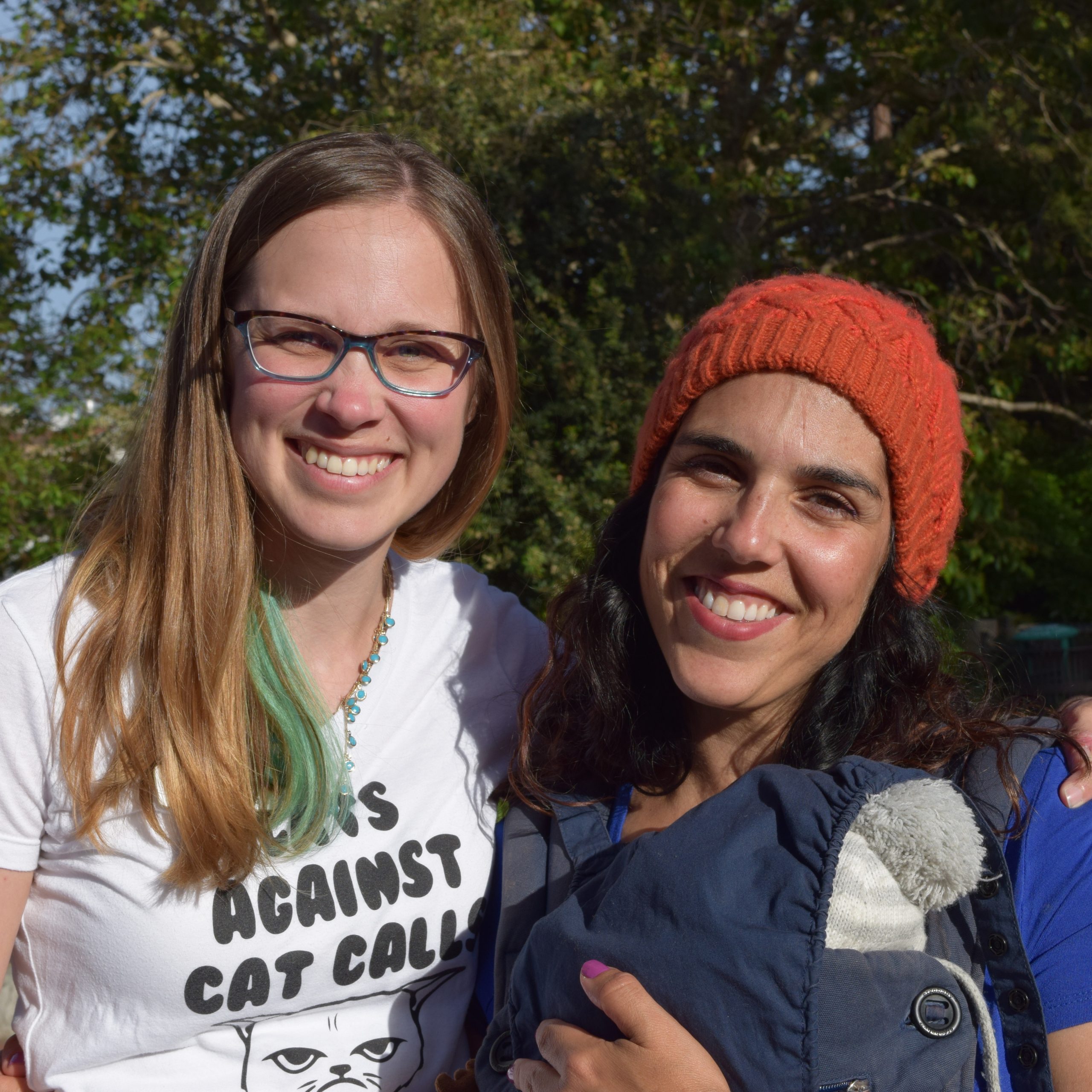A woman in a "Cats Against Catcalls" shirt and a woman in a red beanie and holding a baby smile outdoors