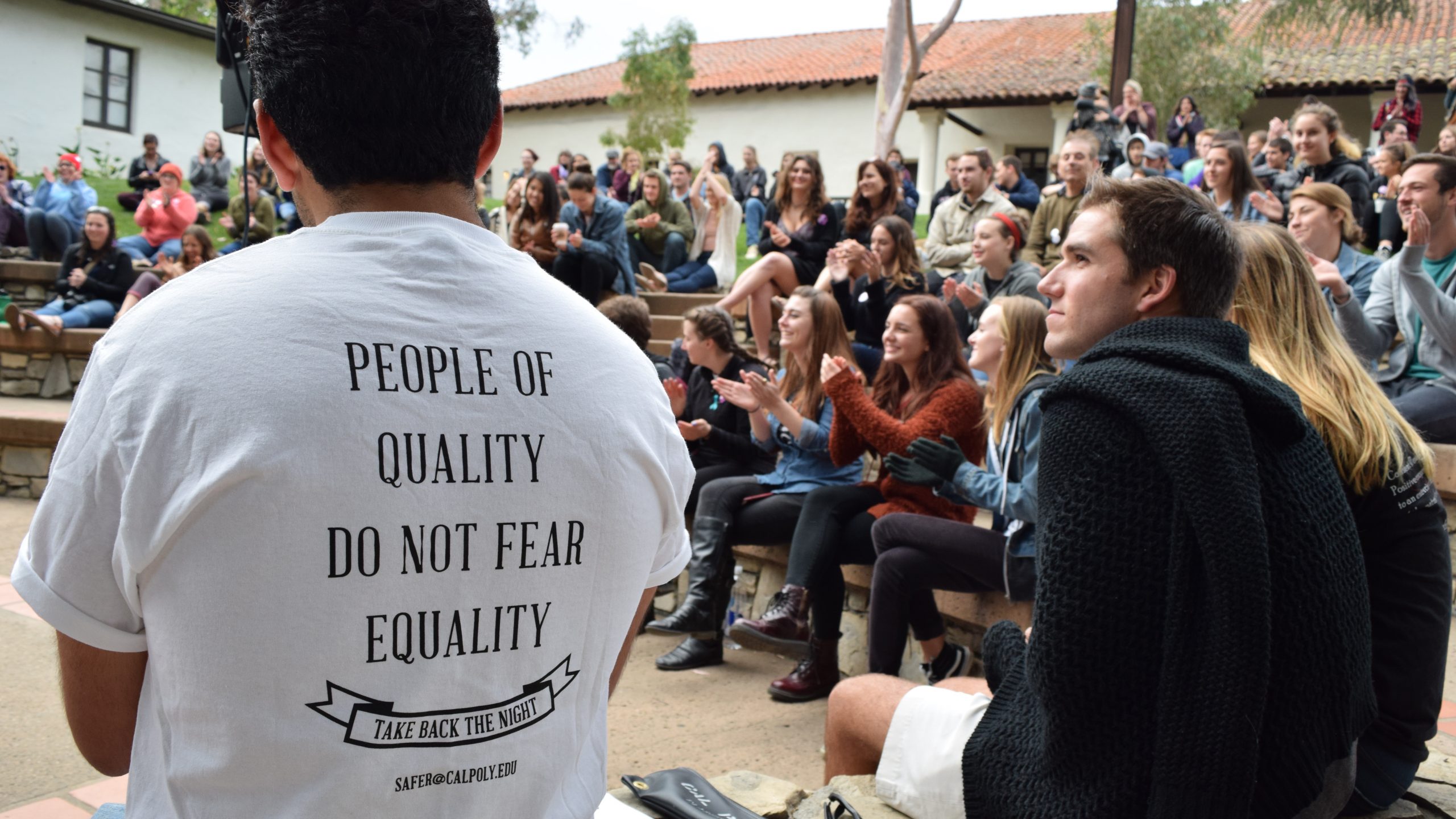 A large group of students applaud a speaker at San Luis Obispo's Mission Plaza. A young man in the foreground wears a shirt reading "People of quality do not fear equality"