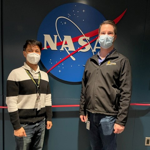 Two men in masks stand in front of a wall with the NASA logo