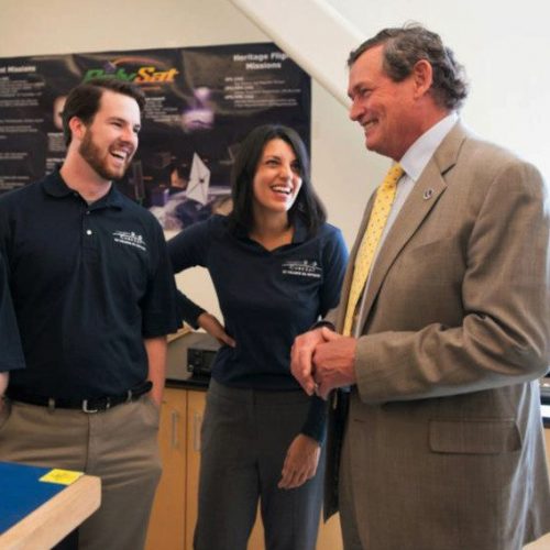 Two students in black polo shirts stand with an older man in a suit, smiling