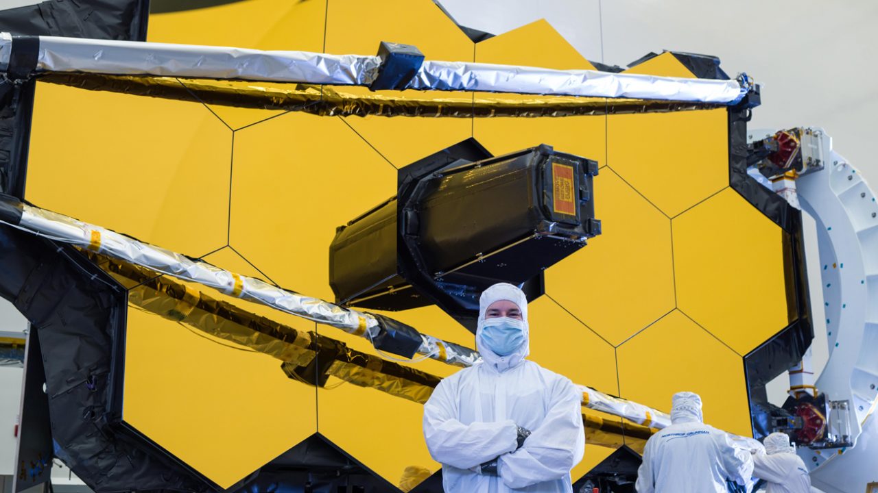 A person wearing a white plastic suit and mask stands with arms crossed in front of the yellow panels of the James Webb telescope