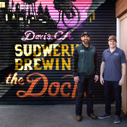 Ryan Fry and Trent Yackzan stand in front of a colorful mural that says 'Davis, CA Sudwerk Brewing the Dock'