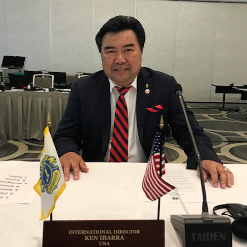 Ken Ibarra sits at a desk with a name plate, flags and a microphone