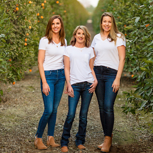 Three people wearing white t-shirts and jeans stand in a citrus orchard