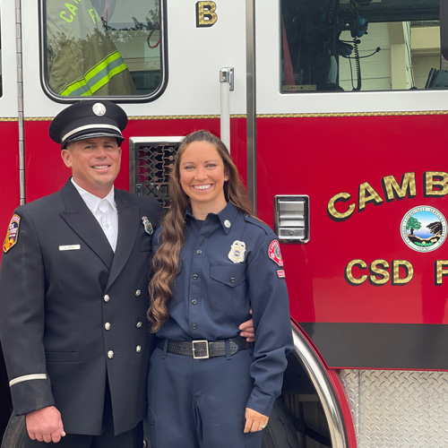 Kayla Graves stands in uniform in front of a red fire truck with another fire department employee