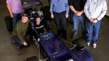The leader of Cal Poly Racing sits in a blue Formula 1 car with the number 20 surrounded by seven alumni