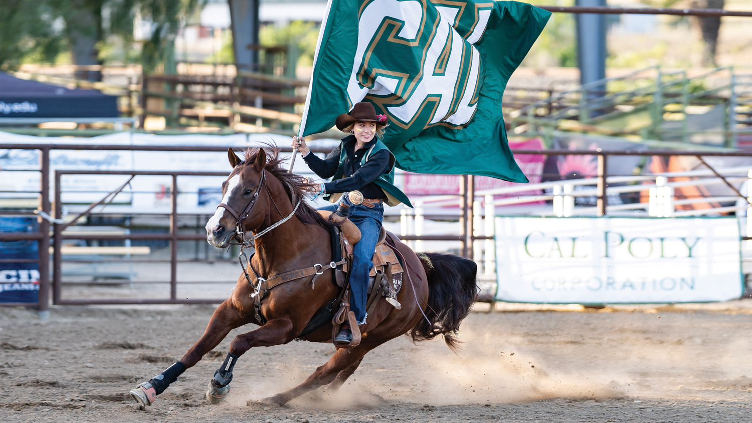 A female student in jeans and a cowboy hat holds a green flag that says "Cal" while riding a brown horse in the Cotton Rosser Rodeo Complex