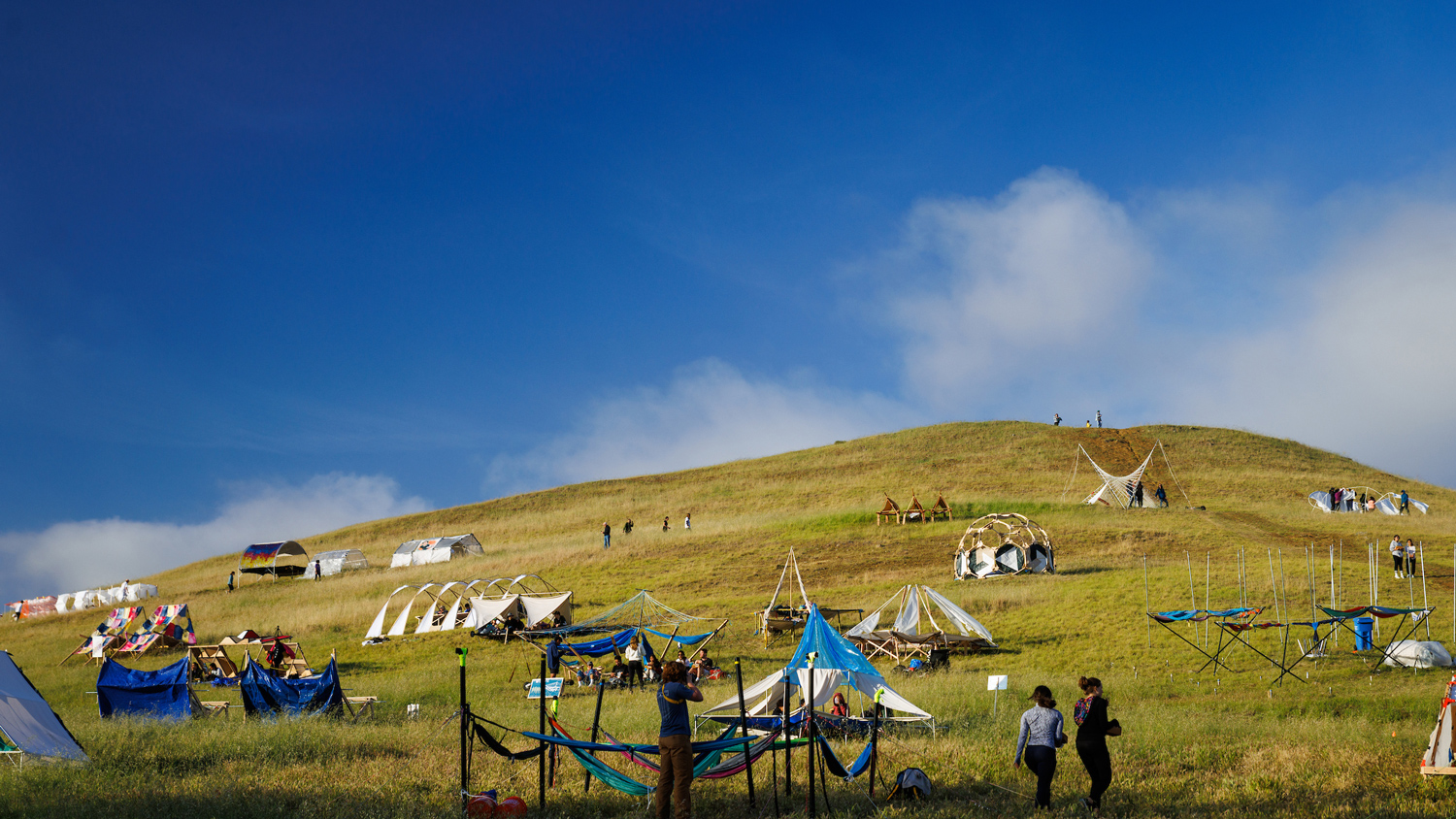 Student-made structures dot the green hillsides of Poly Canyon under blue sky