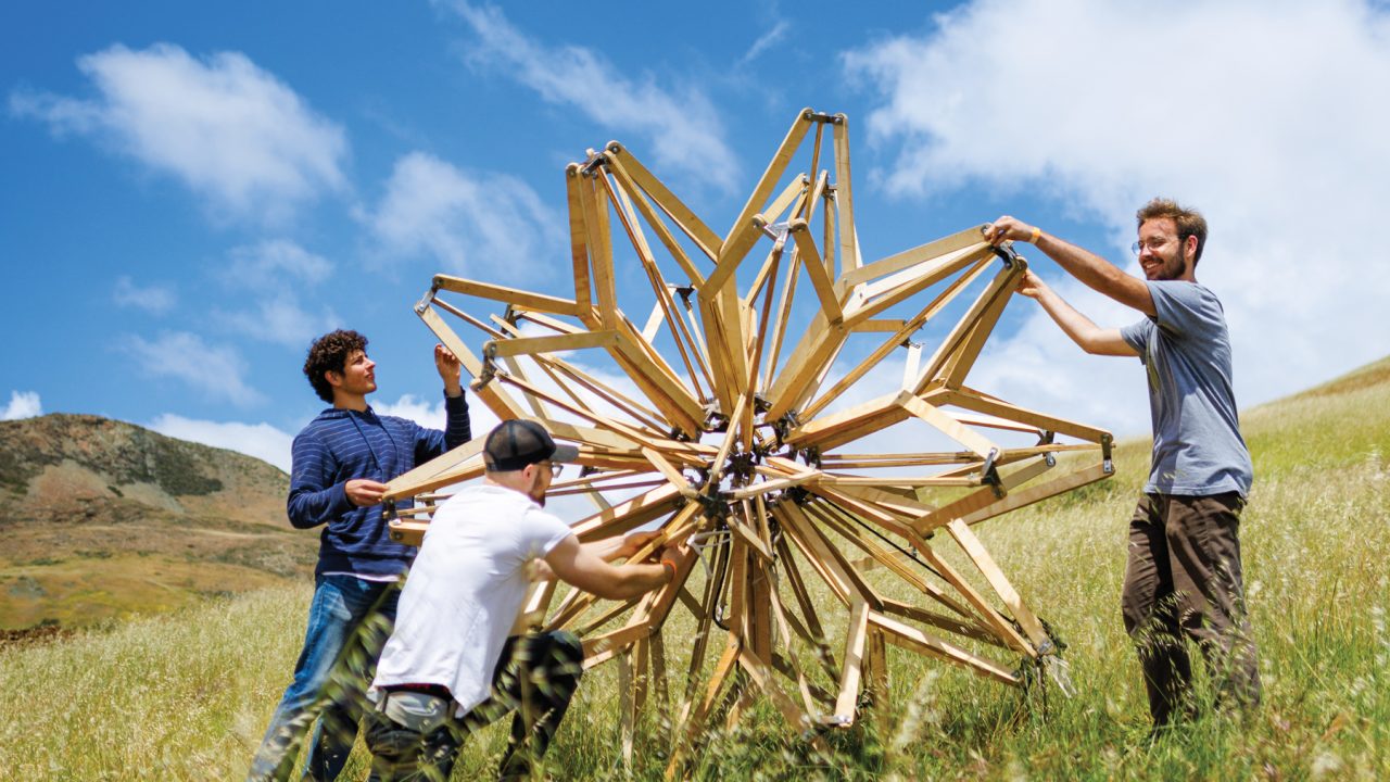 Three students install a lifesize Hoberman Sphere on a grassy hillside under blue skies