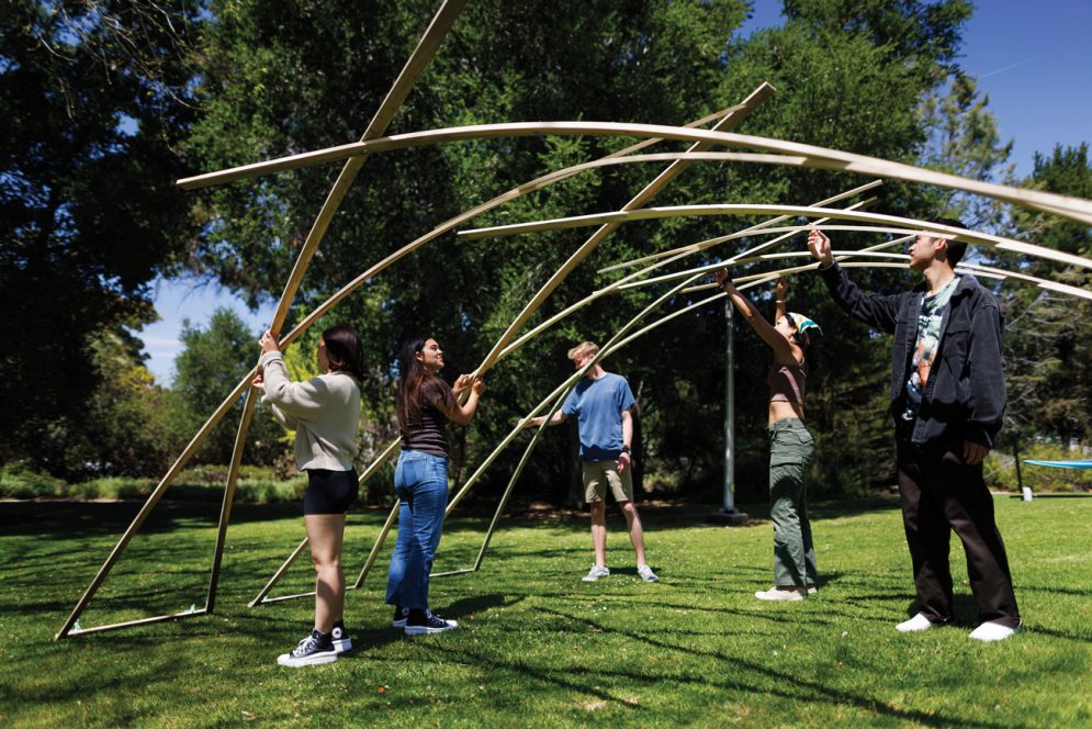 Five students hoist up a wooden lattice structure on a lawn