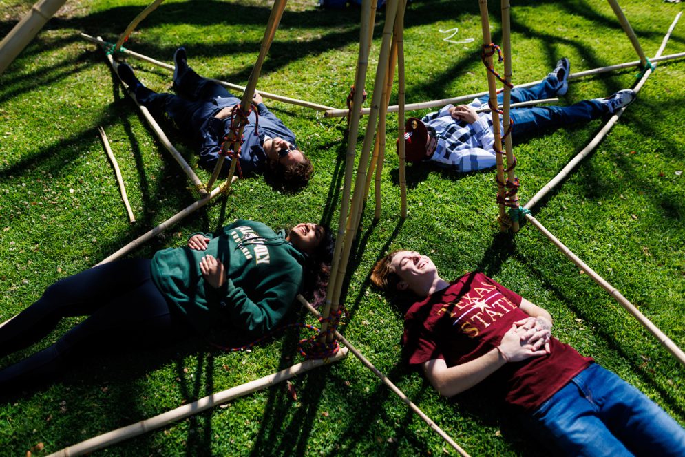 Four students lay on green grass in the bamboo frame of their structure
