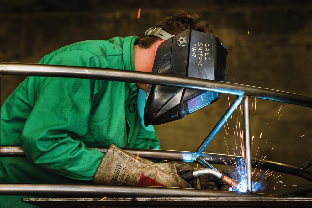 A student in a green coat and face shield welds on steel posts, yielding sparks. 