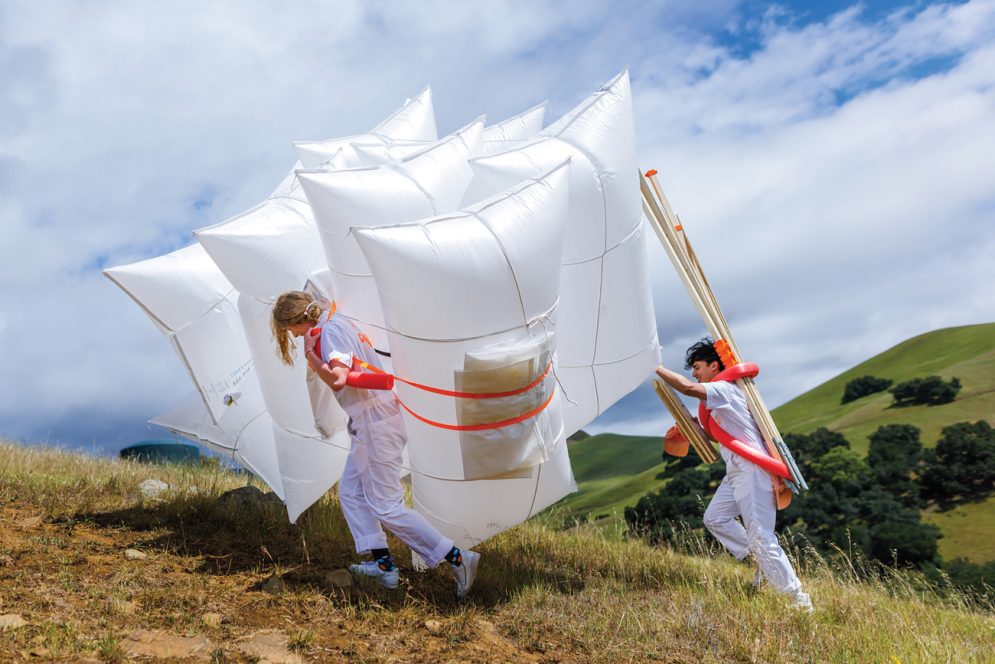Two people wearing white carry inflated white bags and posts up a steep hill