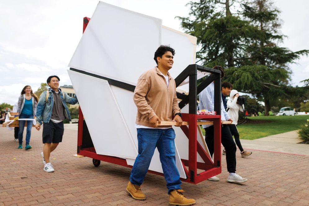 Five students smile while rolling a white, black and red experimental structure over bricks near Dexter Lawn