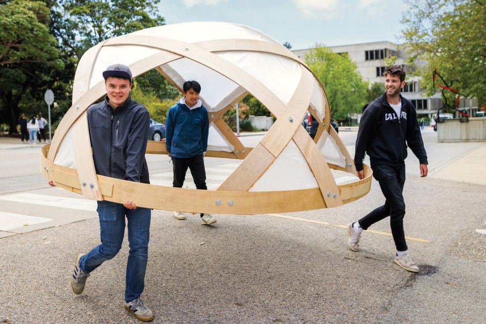 Three students carry a wooden dome near Kennedy Library