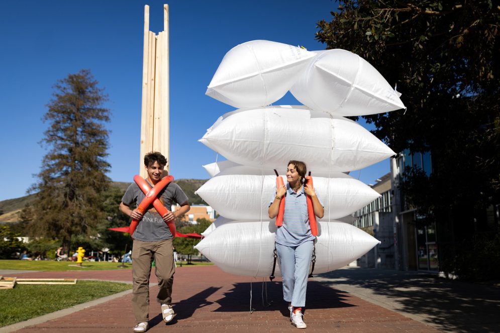 Two students walk with large inflated white bags and tall wooden posts strapped to their backs