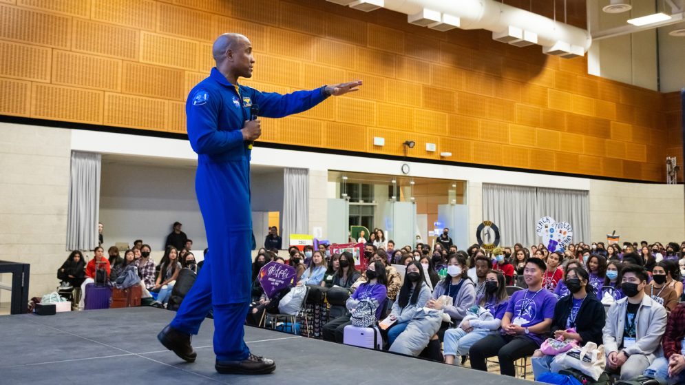 Captain Victor Glover wears a blue flight suit while addressing a crowd of students seated in an auditorium