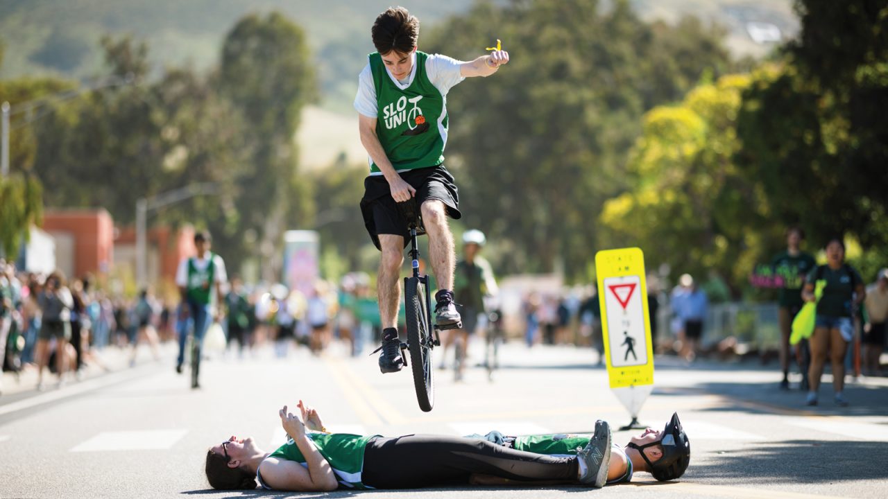 A person riding a unicycle jumps over two people laying on the pavement during an Open House parade at Cal Poly