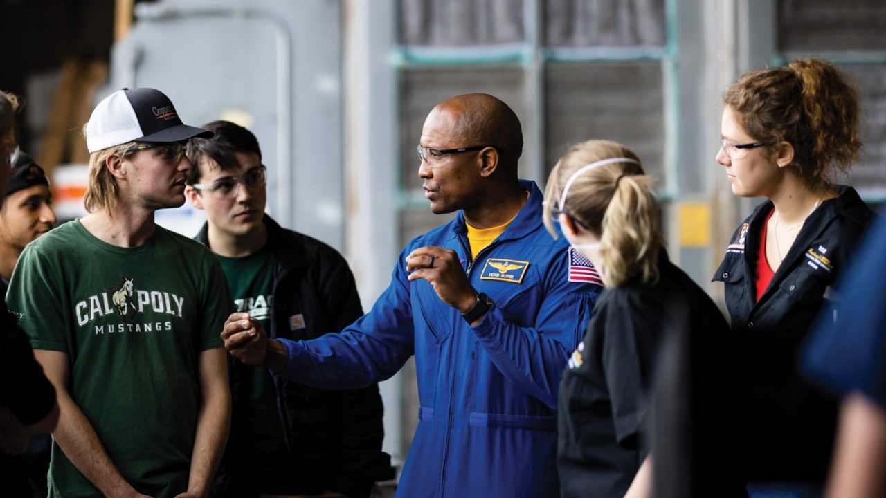 Astronaut Victor Glover wears a blue flight suit while talking to students in a Cal Poly engineering lab