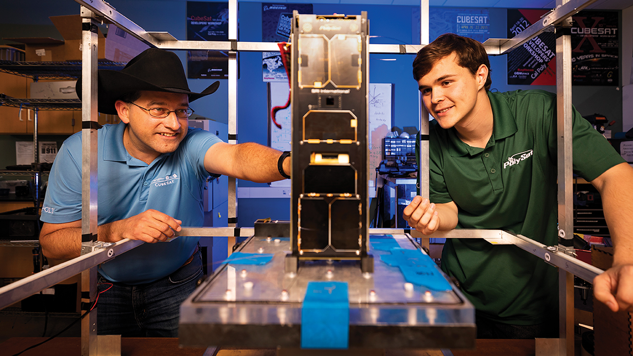 A professor in a blue shirt and cowboy hat and a student in a green polo examine a miniature satellite in a lab