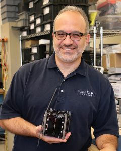 A professor in a black polo holds a miniature satellite in his hand