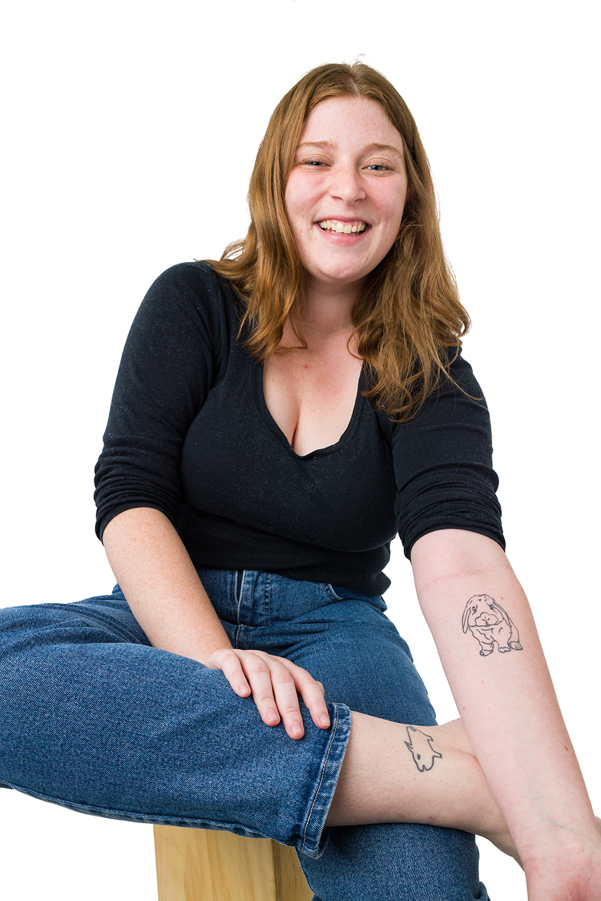 A young woman in a black shirt and jeans smiles as she shows off a rabbit tattoo on her arm and a pig tattoo on her ankle