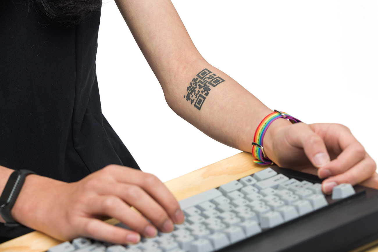 A close-up of two arms poised over a keyboard, one tattooed with a QR code