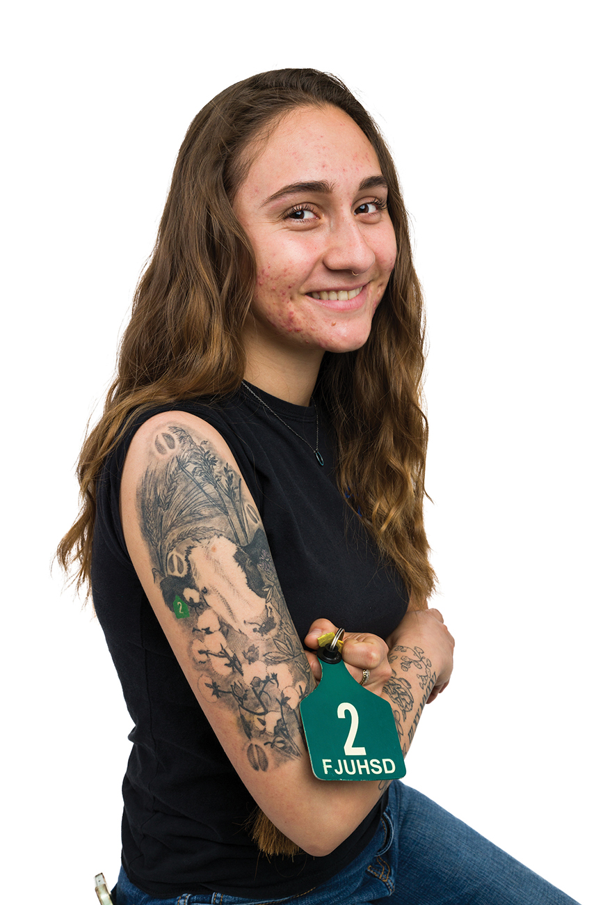 A young woman holds a green cattle tag while rolling up her sleeve to show a tattoo of a cow