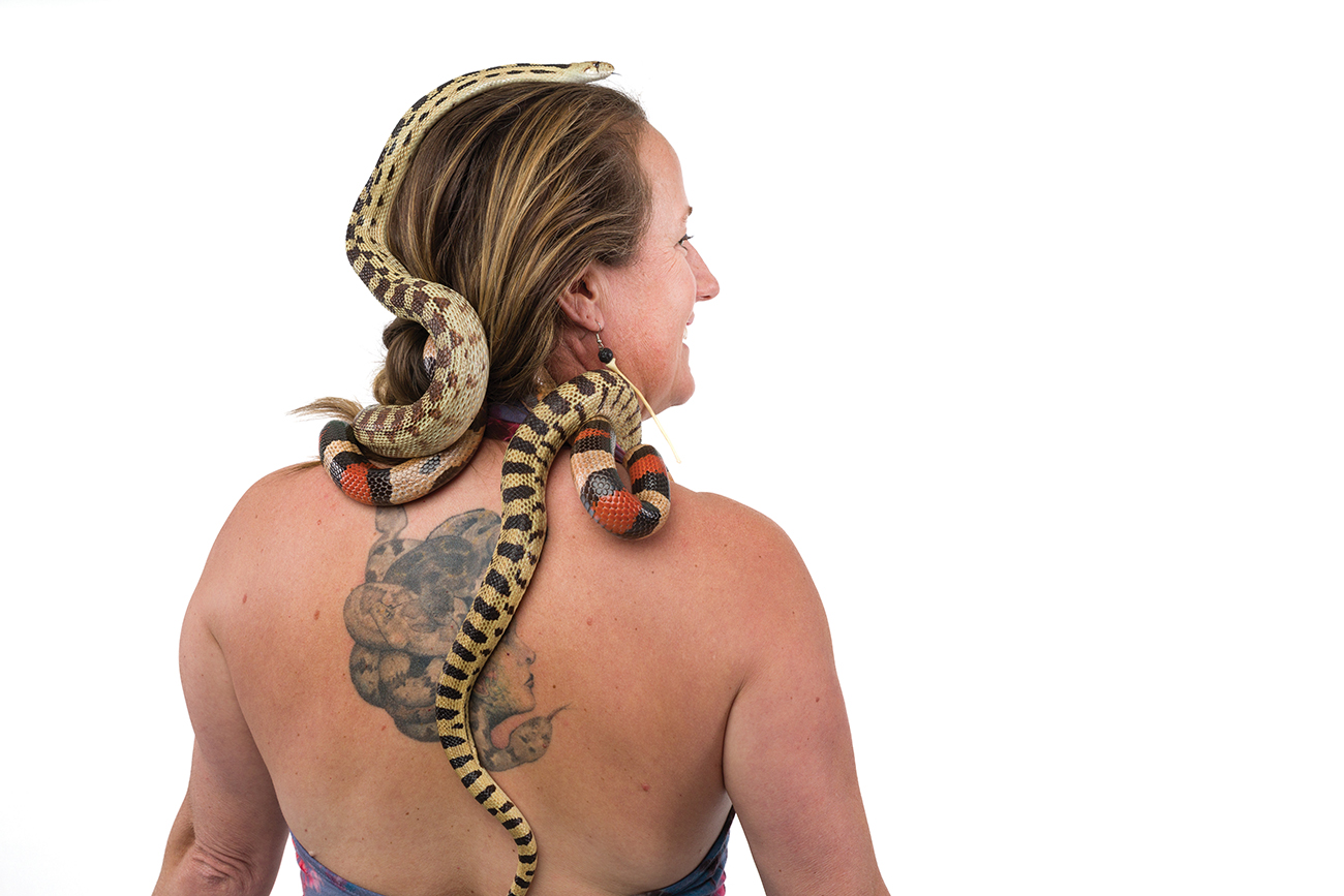 A woman shows off a tattoo of Medusa on her upper back while live snakes drape on her head and shoulders