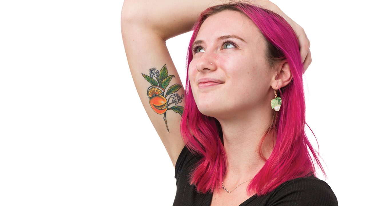 A young woman with pink hair raises her arm to display a brightly colored tattoo of oranges, a branch and orange blossoms