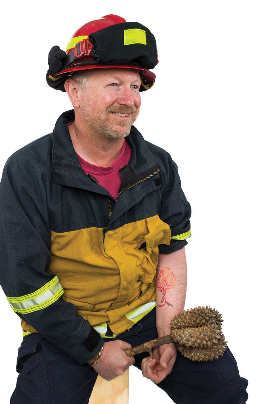 A man in a firefighter's coat and helmet holds a branch of pine cones and displays a tattoo of a tree made of flames on his arm