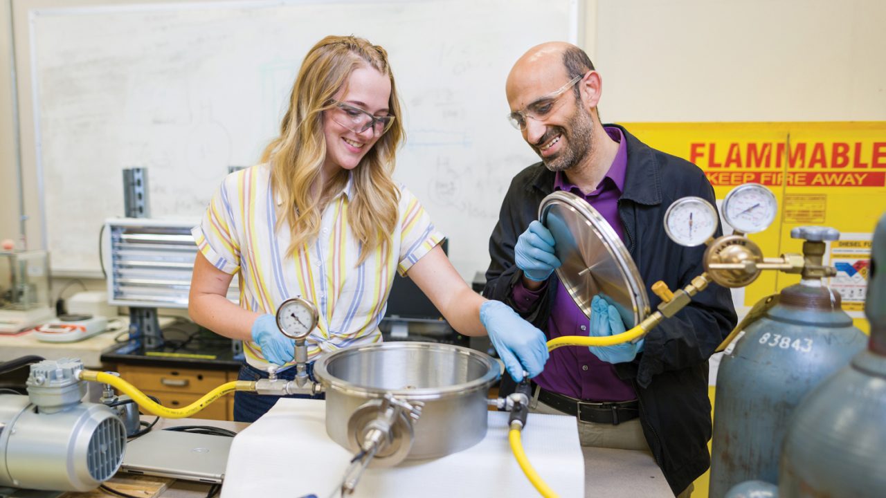 A teacher and student wear gloves and goggles while working in an engineering lab