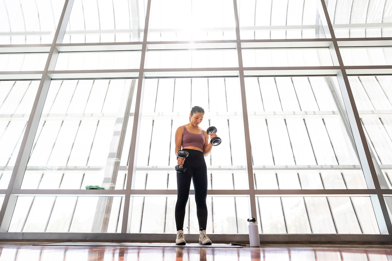 A young woman curls dumbbells in front of a large wall of windows