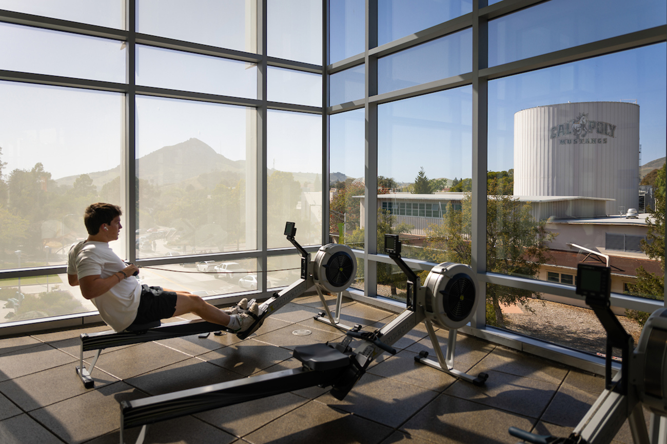 A young man exercises on a rowing machine in front of a wall of windows overlooking campus and Bishop Peak
