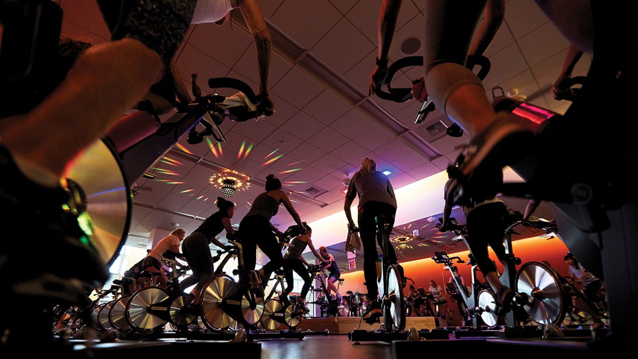A low-angle view of a cycling class in a dark room with colorful lights
