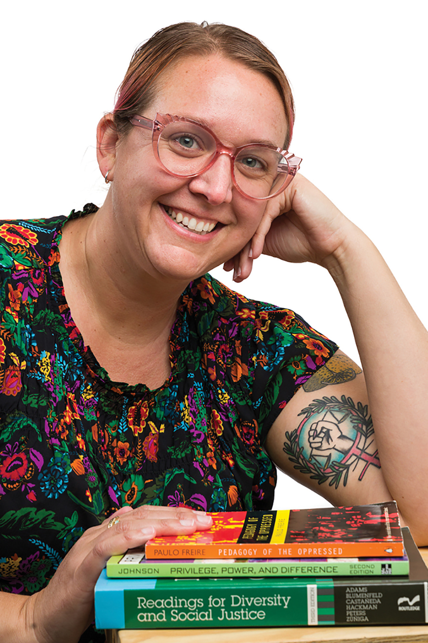 A woman leans on a desk in front of a pile of books, showing off a brightly colored arm tattoo