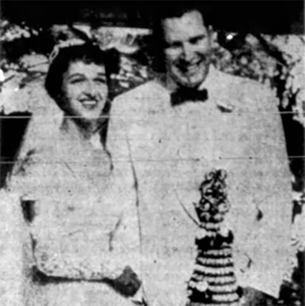 A young Virginia and John, in wedding attire, stand in front of their wedding cake