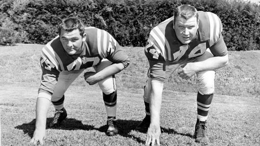 A young John Madden in a football uniform lines up in a three point stance with Cal Poly teammate Pat Lovell