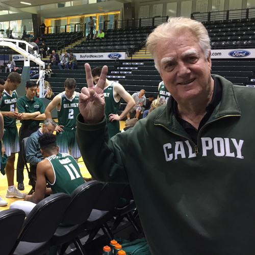 Jeffrey Wilson wears a green Cal Poly sweater and gives a peace sign while watching the Mustang Men's Basketball Team huddle