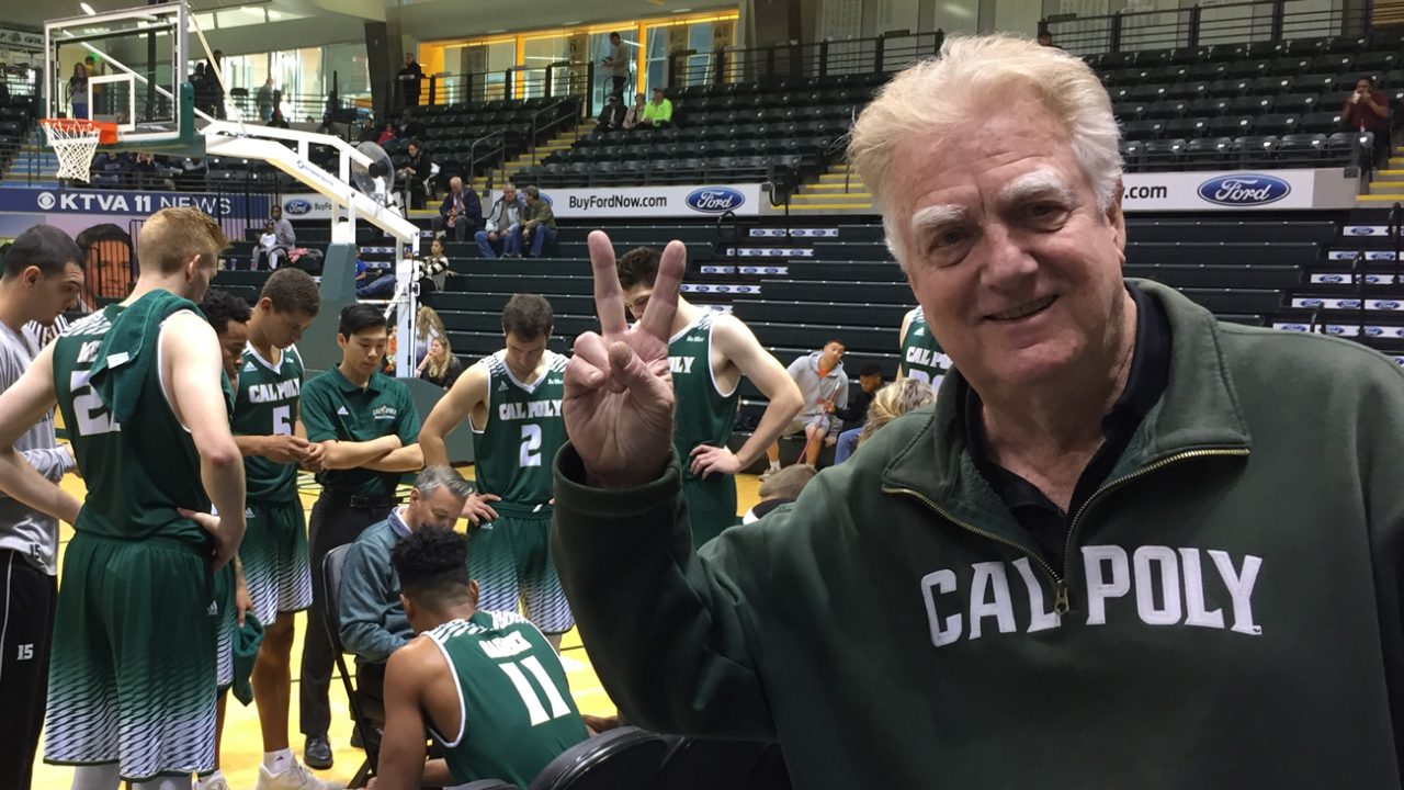Jeffrey Wilson wears a green Cal Poly sweater and gives a peace sign while watching the Mustang Men's Basketball Team huddle