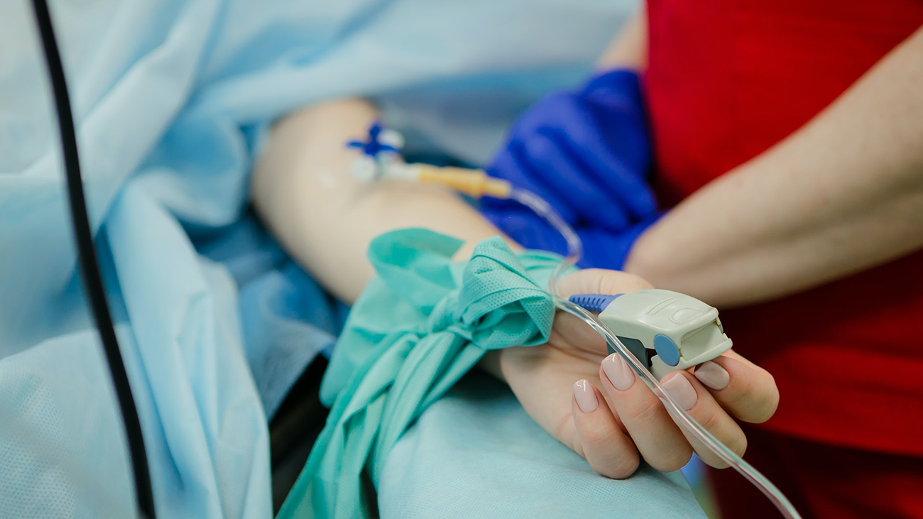 A patient's arm, with an IV drip and a heart monitor attached, protrudes from under a sterile drape on an operating table