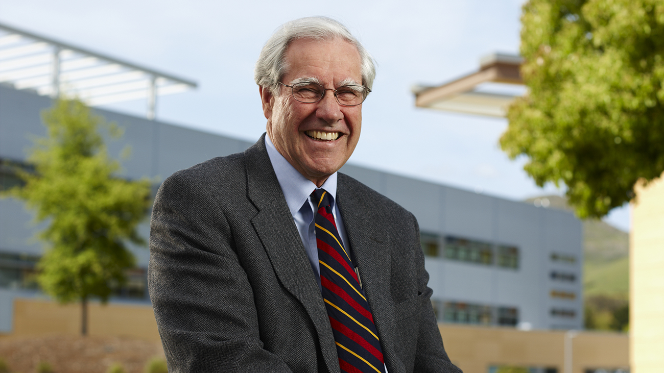 Former President Warren Baker grins in a suit and tie while sitting in front of the Engineering Plaza