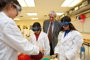 Warren Baker watches students in lab coats and goggles perform a scientific experiment.