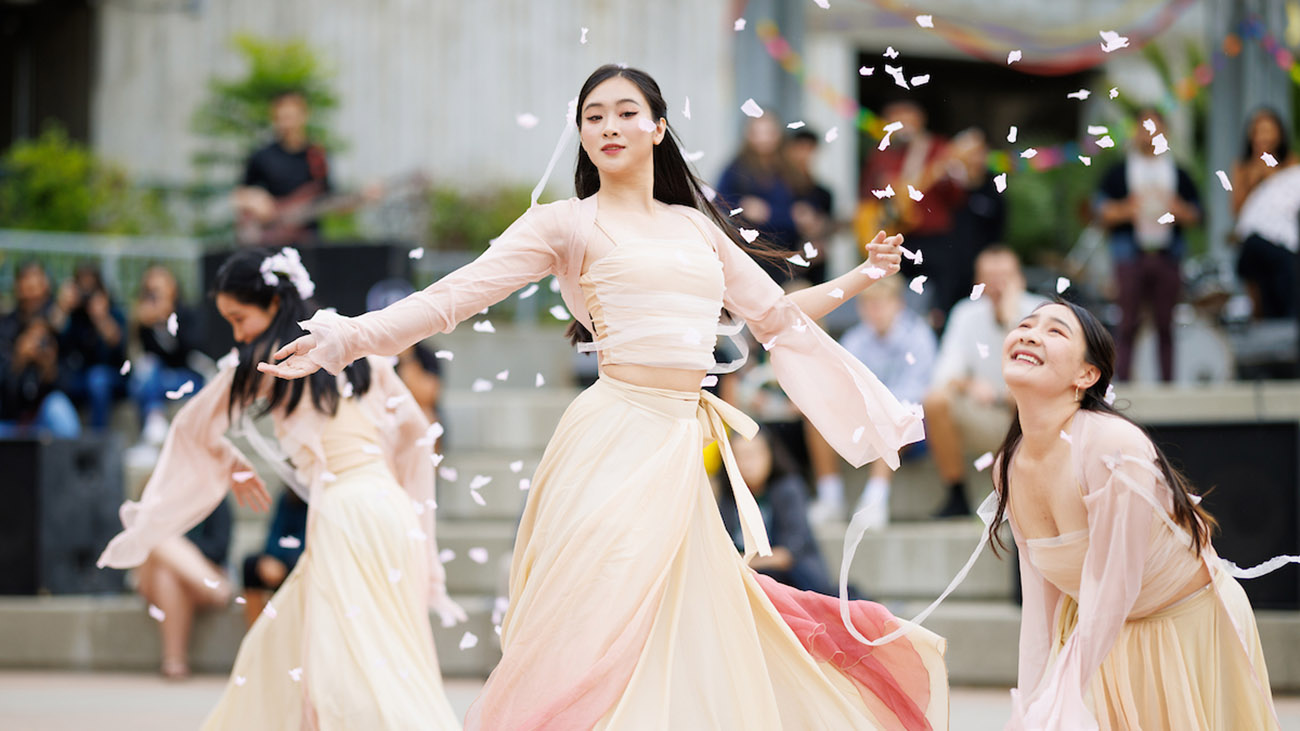 Three young women in flowing white and pink gowns strike dramatic dance poses as flower petals swirl around them.