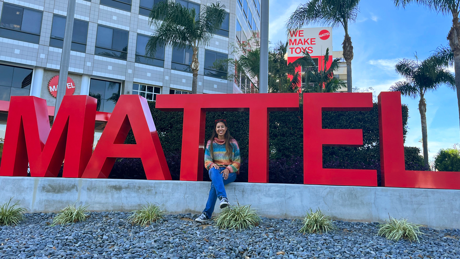 Kristen Sanzari sits on a red sign for Mattel in front of an office building