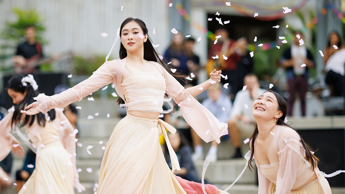 Three young women in flowing pink and white gowns dance dramatically while flower petals swirl around them
